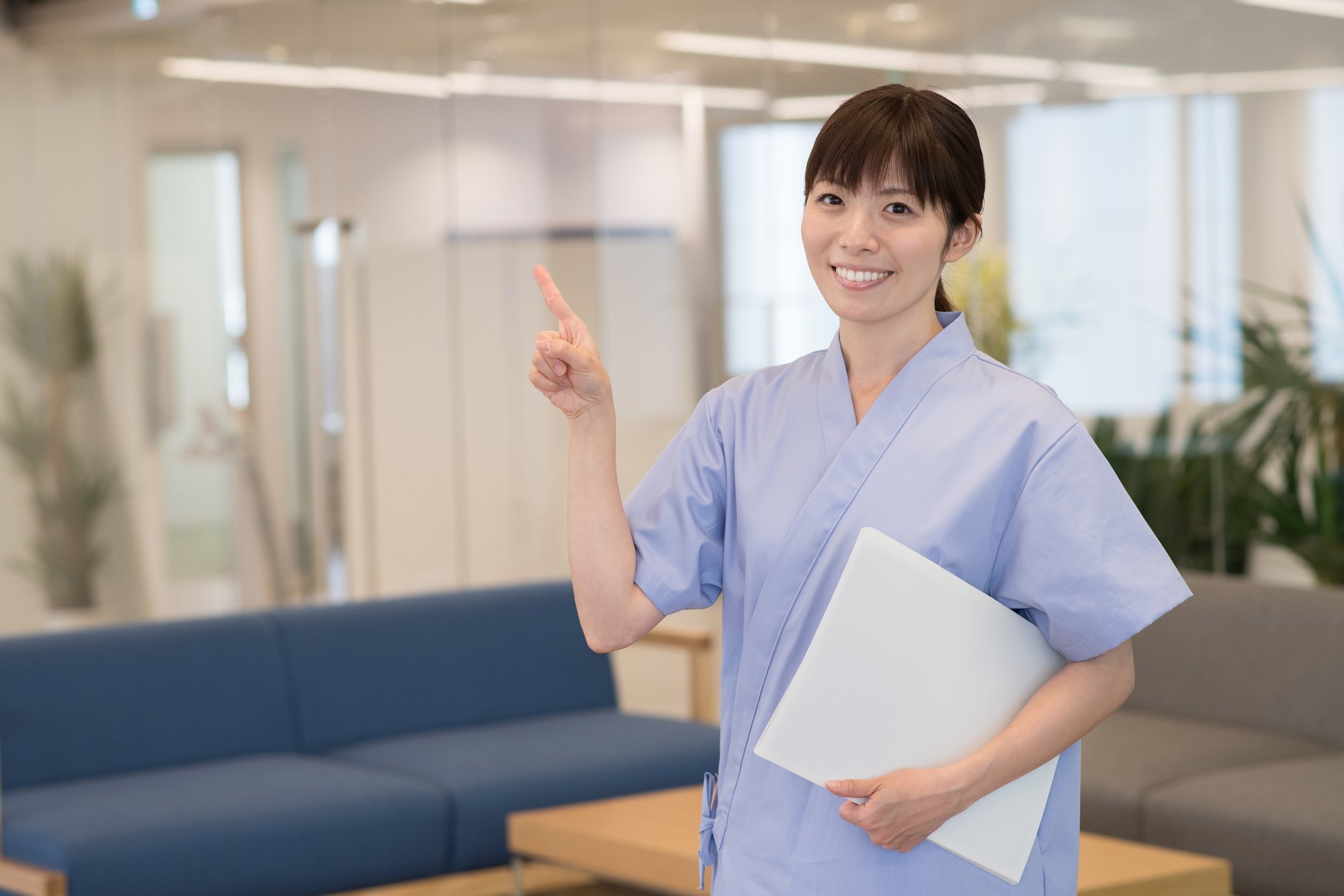 資料を片手に持ち、人さし指を上に向ける検査着姿の女性